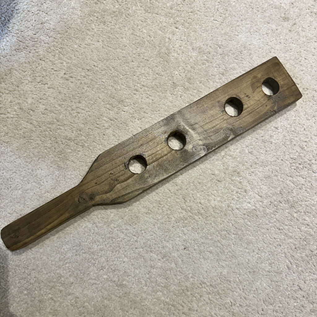 Handcrafted wooden paddle with four holes on a light-colored carpet.